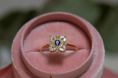 Anillo de plata 925 con baño de oro 18k y piedras centrales en forma de flor de zirconias blancas y azul exhibido en caja de anillo rosa en Guatemala