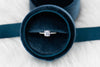 Anillo de plata 925 ajustable con zirconia central grande y zirconias pequeñas incrustadas en la banda exhibido de frente dentro de caja de anillo Negra con azul