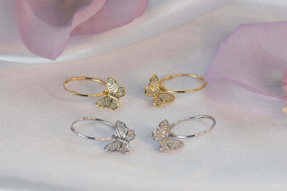 un par de argollas en plata 925 con mariposa colgante con zirconias y otro par de argollas en plata 925 con baño en oro 18k con mariposa colgante con zirconias exhibido sobre superficie blanca