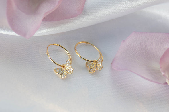 un par de argollas en plata 925 con baño en oro 18k con mariposa colgante con zirconias exhibido sobre superficie blanca