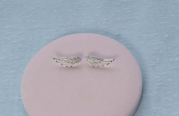 Aretes de plata 925 en forma de alas de ángel exhibidos sobre superficie rosa