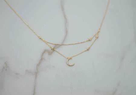 Collar doble de cadenas con pendiente de luna con incrustaciones en zirconias y estrellas en baño de oro 18k sobre superficie de mármol